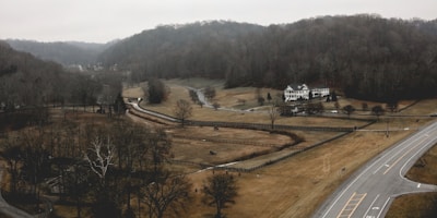 A rural landscape featuring a large two-story white house surrounded by expansive fields with sparse trees. A winding road cuts through the scene, leading past the house and into the distance. Hills densely covered with bare trees form the backdrop, under a gray, overcast sky.
