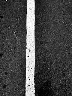 A vertical white line on a dark asphalt surface, showing some wear and texture differences on the pavement.