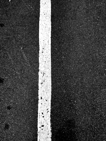 A vertical white line on a dark asphalt surface, showing some wear and texture differences on the pavement.