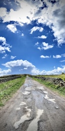 A rural, unpaved road stretches into the distance, bordered by green grassy fields and stone walls. The sky is expansive and vibrant with large, fluffy white clouds scattered across a deep blue background. Puddles and patches of wet soil are visible on the road, suggesting recent rain.