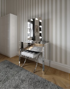 A modern vanity setup featuring a mirrored table with a lighted mirror and a tufted stool beneath it. The room has striped wallpaper and a large wardrobe. The floor is wooden with a plush gray rug, and a window with curtains is visible beside a framed wall decoration.