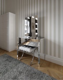 A modern vanity setup featuring a mirrored table with a lighted mirror and a tufted stool beneath it. The room has striped wallpaper and a large wardrobe. The floor is wooden with a plush gray rug, and a window with curtains is visible beside a framed wall decoration.