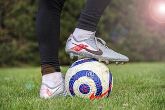 A person is wearing soccer cleats and standing on a soccer ball on a grassy field. The cleats are white with red and black accents, and the ball has a blue and red design. There is a lens flare effect in the top right corner, adding a bright spot.