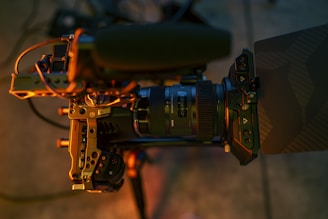 A close-up view of a professional video camera setup, featuring a large lens with brand labels, a microphone attached to the top, various buttons and adjustment knobs, all mounted on a tripod. The lighting casts warm, golden tones across the equipment.