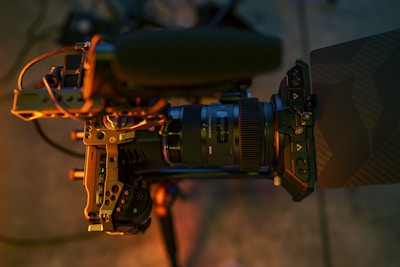 A close-up view of a professional video camera setup, featuring a large lens with brand labels, a microphone attached to the top, various buttons and adjustment knobs, all mounted on a tripod. The lighting casts warm, golden tones across the equipment.
