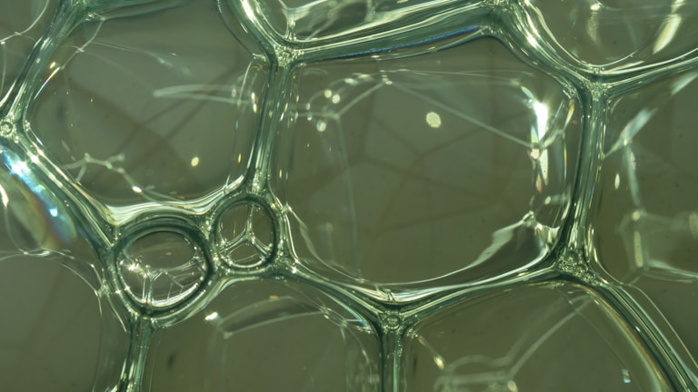 A close-up view of transparent soap bubbles clustered together, creating an intricate network of hexagonal and irregular shapes. The bubbles reflect light, casting subtle rainbow colors and providing a sense of fragility and delicacy.