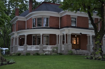 A large, stately brick house with a wraparound porch, supported by white columns. The house features several windows with curtains, a dark roof, and lush greenery surrounding it. The evening light casts a warm glow, creating a serene atmosphere.