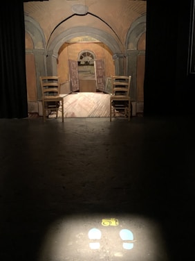 A room features an artistic backdrop resembling a doorway leading to another room, with artistic brush strokes on the walls and floor. Two wooden chairs are positioned symmetrically on each side. A spotlight on the floor highlights a drawing of a camera and two footprints, suggesting a photo opportunity.