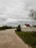 A rural scene with a dirt road curving to the left, surrounded by grass and sparse trees under an overcast sky. A small house with a red roof and white walls is situated on the right, behind a concrete fence.