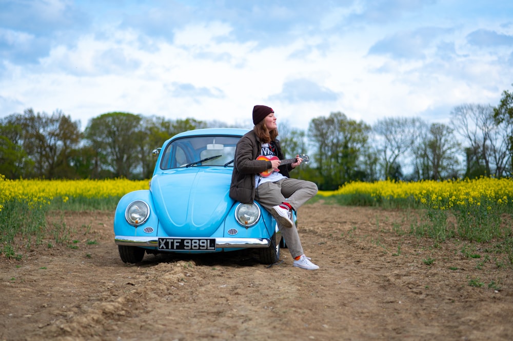 man in black jacket and gray pants sitting on blue volkswagen beetle