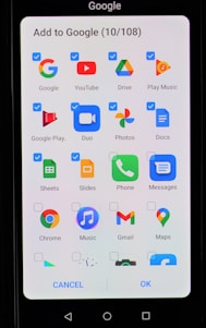 A smartphone screen displaying an interface to add applications to Google. The interface includes multiple app icons such as Google, YouTube, Drive, Play Music, Google Play, Duo, Photos, Docs, Sheets, Slides, Phone, Messages, Chrome, Music, Gmail, and Maps. Each app icon has a checkbox next to it, some of which are checked.