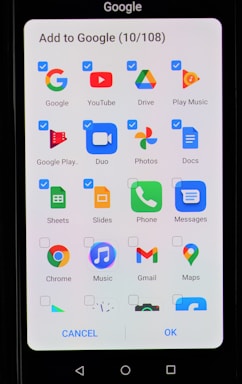 A smartphone screen displaying an interface to add applications to Google. The interface includes multiple app icons such as Google, YouTube, Drive, Play Music, Google Play, Duo, Photos, Docs, Sheets, Slides, Phone, Messages, Chrome, Music, Gmail, and Maps. Each app icon has a checkbox next to it, some of which are checked.