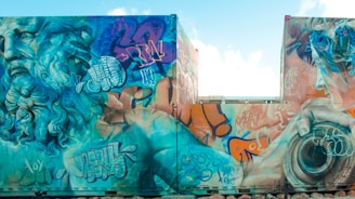 A vibrant mural featuring a large, detailed depiction of a classical figure with long, flowing hair and beard. The artwork is filled with various graffiti tags and abstract elements surrounding the figure, leading to a blend of traditional and urban art styles. The colors are bold and contrasting, with a strong emphasis on blues, oranges, and greens.