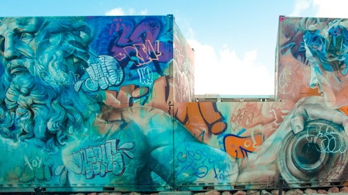 A vibrant mural featuring a large, detailed depiction of a classical figure with long, flowing hair and beard. The artwork is filled with various graffiti tags and abstract elements surrounding the figure, leading to a blend of traditional and urban art styles. The colors are bold and contrasting, with a strong emphasis on blues, oranges, and greens.