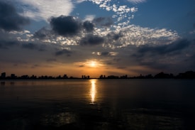The image captures a serene sunset over a city skyline. The sun is partially hidden by scattered clouds, creating a warm glow across the sky. Reflections of the sun can be seen shimmering across the calm water in the foreground.