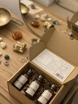 A cardboard box containing three bottles labeled 'SIPS Brain' is open on a wooden table. The box is packed with crinkle paper for protection. Surrounding the box on the table are various items including stones, a decorative bowl, amber-colored stones, and jewelry pieces such as rings. A mirror and a decorative object are also visible on the table.