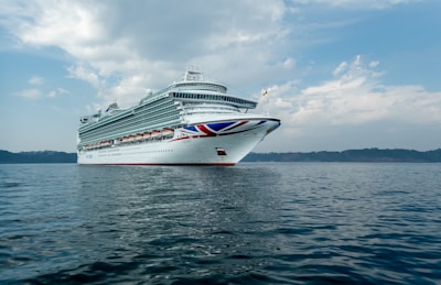A large cruise ship is sailing on calm waters under a partly cloudy sky. The ship has multiple decks with numerous windows and is decorated with a red, white, and blue design on its bow.