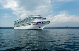 A large cruise ship is sailing on calm waters under a partly cloudy sky. The ship has multiple decks with numerous windows and is decorated with a red, white, and blue design on its bow.