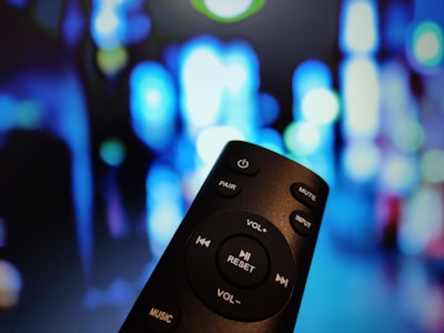 A remote control is in focus in the foreground, featuring buttons for power, volume, and other functions. The background is filled with vibrant bokeh lights, predominantly in shades of blue and green, creating an abstract and dynamic atmosphere.