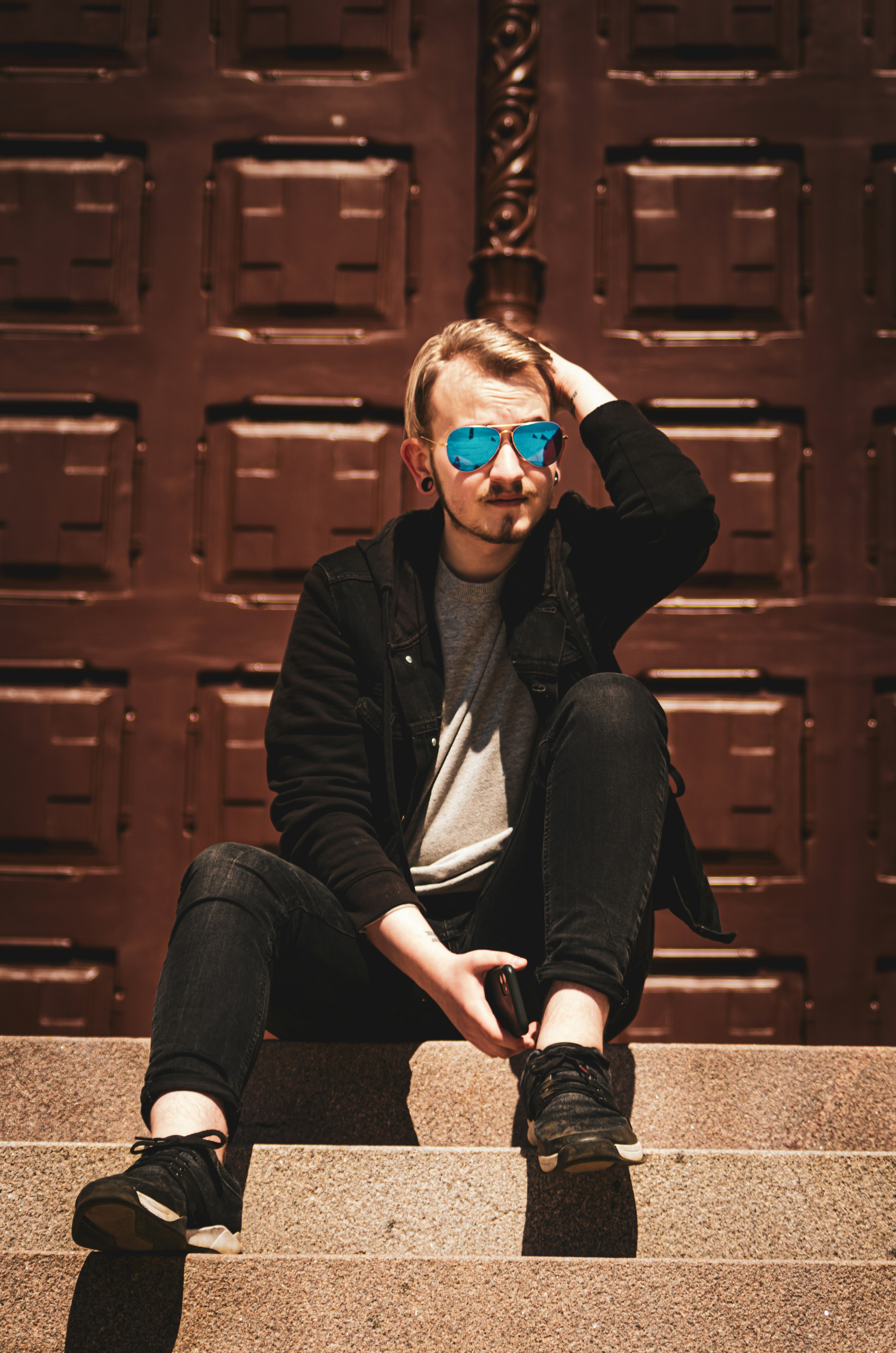 woman in black jacket and blue denim jeans wearing blue sunglasses sitting on brown wooden bench