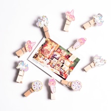A circular arrangement of wooden clips decorated with cartoon unicorns, cupcakes, and donuts. In the center, a photograph displaying a collage of framed art and vintage items is attached. The background is a plain white surface.