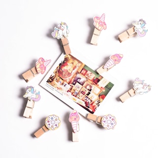 A circular arrangement of wooden clips decorated with cartoon unicorns, cupcakes, and donuts. In the center, a photograph displaying a collage of framed art and vintage items is attached. The background is a plain white surface.