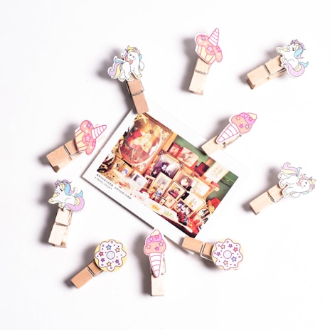 A circular arrangement of wooden clips decorated with cartoon unicorns, cupcakes, and donuts. In the center, a photograph displaying a collage of framed art and vintage items is attached. The background is a plain white surface.