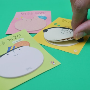 Three colorful sticky note pads featuring cartoonish animal faces on a bright green background. A hand is peeling back one of the sticky notes, revealing additional layers underneath.