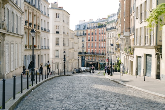 A cobblestone street lined with historic Parisian buildings, featuring multiple floors with balconies and several street lamps. People are walking along the street, and the sky is bright and clear, suggesting a sunny day. The scene conveys a classic European city atmosphere.