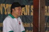 A man wearing a white and green polo shirt with an emblem, and a black cap, is speaking into a microphone. The background is a blurred banner with large yellow text.
