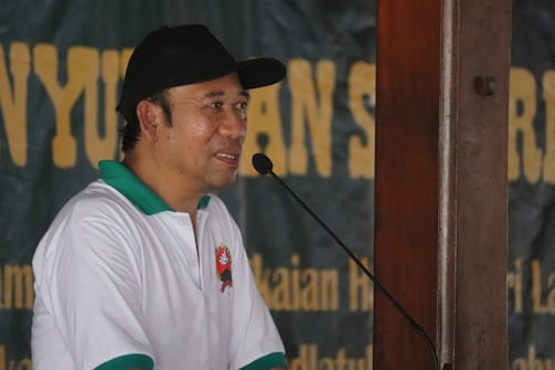 A man wearing a white and green polo shirt with an emblem, and a black cap, is speaking into a microphone. The background is a blurred banner with large yellow text.