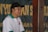 A man wearing a white and green polo shirt with an emblem, and a black cap, is speaking into a microphone. The background is a blurred banner with large yellow text.