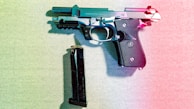 A handgun is placed on a textured surface with a magazine lying next to it. The image is lit with a gradient light effect, combining blue and red hues that cast colorful shadows. The handgun has a black grip with metallic accents and is resting horizontally.