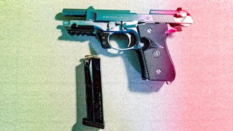 A handgun is placed on a textured surface with a magazine lying next to it. The image is lit with a gradient light effect, combining blue and red hues that cast colorful shadows. The handgun has a black grip with metallic accents and is resting horizontally.