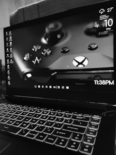 A laptop displaying a close-up image of a gaming controller on its screen. The room temperature is shown as 27°C with thunderstorm predictions. The date displayed is May 10 and the time is 11:38 PM. The laptop's keyboard is illuminated, emphasizing its design in a dimly lit environment.
