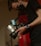 A person wearing a black mask is holding a professional camera with a label that reads 'BEN COLLINS BLACKMAGIC 1'. The person is wearing a black short-sleeved shirt and a blue wristband. The environment appears dimly lit, creating a focused and professional atmosphere.