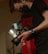 A person wearing a black mask is holding a professional camera with a label that reads 'BEN COLLINS BLACKMAGIC 1'. The person is wearing a black short-sleeved shirt and a blue wristband. The environment appears dimly lit, creating a focused and professional atmosphere.