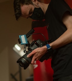 A person wearing a black mask is holding a professional camera with a label that reads 'BEN COLLINS BLACKMAGIC 1'. The person is wearing a black short-sleeved shirt and a blue wristband. The environment appears dimly lit, creating a focused and professional atmosphere.