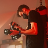 A person wearing a black face mask and a black t-shirt is holding a professional video camera. The setting appears to be dimly lit with a red-orange hue, suggesting a possibly professional or creative environment.