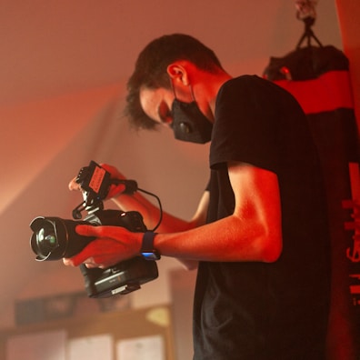 A person wearing a black face mask and a black t-shirt is holding a professional video camera. The setting appears to be dimly lit with a red-orange hue, suggesting a possibly professional or creative environment.