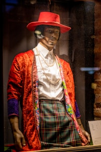 A mannequin dressed in a colorful and eclectic outfit, featuring a vibrant red hat, a red textured jacket with floral-lined edges, a white dress shirt, and plaid trousers. The ensemble is accessorized with layered gold necklaces in what appears to be a store display.