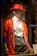 A mannequin dressed in a colorful and eclectic outfit, featuring a vibrant red hat, a red textured jacket with floral-lined edges, a white dress shirt, and plaid trousers. The ensemble is accessorized with layered gold necklaces in what appears to be a store display.