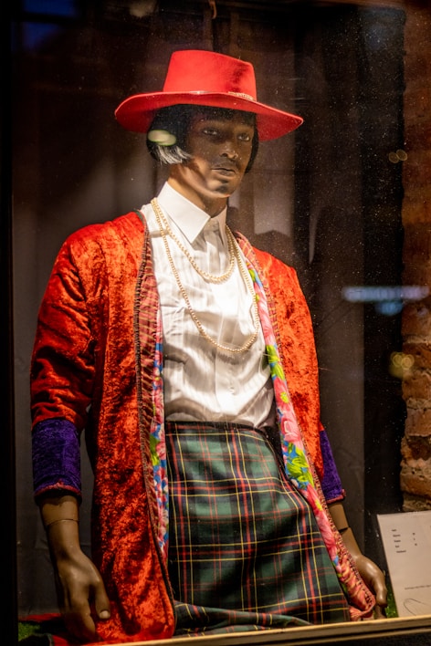 A mannequin dressed in a colorful and eclectic outfit, featuring a vibrant red hat, a red textured jacket with floral-lined edges, a white dress shirt, and plaid trousers. The ensemble is accessorized with layered gold necklaces in what appears to be a store display.