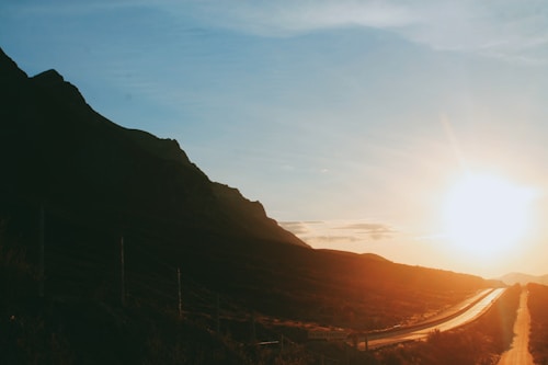 A road curves through a mountainous landscape with a bright sun setting or rising on the horizon, casting a warm golden light. The silhouette of the mountains contrasts with the clear blue sky.