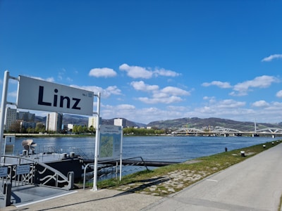A riverside scene with a large sign displaying 'Linz' in the foreground. There are several modern buildings along the riverbank, with a backdrop of green hills and a bridge crossing the river. The sky is bright blue with fluffy white clouds.