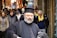 A bearded man wearing traditional black clerical attire is walking through a bustling market street. He appears focused and contemplative. The background is filled with a diverse group of people in casual winter clothing, some with scarves and jackets. Various colorful textiles and merchandise are visible in the stalls lining the street, adding a vibrant atmosphere.