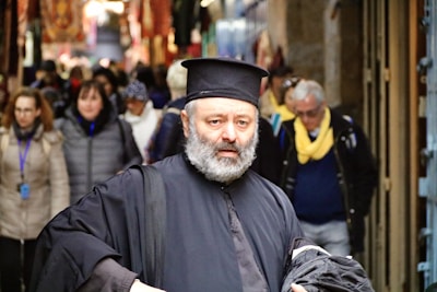 A bearded man wearing traditional black clerical attire is walking through a bustling market street. He appears focused and contemplative. The background is filled with a diverse group of people in casual winter clothing, some with scarves and jackets. Various colorful textiles and merchandise are visible in the stalls lining the street, adding a vibrant atmosphere.