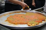 A large metal tray contains a traditional dessert, possibly kunafa, with a golden-orange top layer and sprinkled with green pistachio bits. A person is cutting and serving a piece onto a plate, while stacks of empty plates are visible in the background.