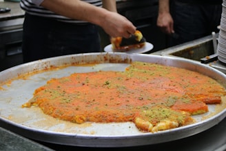 A large metal tray contains a traditional dessert, possibly kunafa, with a golden-orange top layer and sprinkled with green pistachio bits. A person is cutting and serving a piece onto a plate, while stacks of empty plates are visible in the background.