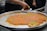 A large metal tray contains a traditional dessert, possibly kunafa, with a golden-orange top layer and sprinkled with green pistachio bits. A person is cutting and serving a piece onto a plate, while stacks of empty plates are visible in the background.